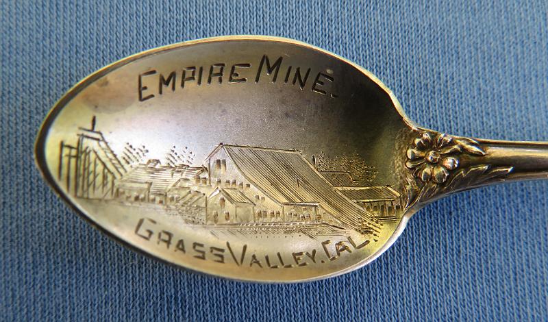 Souvenir Mining Spoon Empire Mine Bowl.JPG - SOUVENIR MINING SPOON EMPIRE MINE GRASS VALLEY CA - Sterling silver souvenir spoon,  features  handle with figural miner behind pan with gold nuggets and pick and shovel, flower with deep relief decoration down the handle, bowl has engraved image of mining scene, marking  is EMPIRE MINE, GRASS VALLEY, CAL., measures 4" in length , reverse marked STERLING with hallmark  of H on a pennant for Mechanics Sterling Company, which was subsidiary of Watson Newell Co. [The city of Grass Valley is the largest city in the western region of Nevada County, California.  Situated at roughly 2,500 feet elevation in the western foothills of the Sierra Nevada mountain range, this historic northern Gold Country city is 57 miles north by northeast  from the state capitol in Sacramento. Grass Valley, which was originally known as Boston Ravine and later officially named Centerville, dates from the California Gold Rush, as does nearby Nevada City. When a post office was established in 1851, it was renamed Grass Valley the following year.   The town incorporated in 1860. Grass Valley is the location of the Empire Mine and North Star Mine, two of the oldest, largest, deepest, longest and richest gold mines in California.  Many of those who came to settle in Grass Valley were tin miners from Cornwall, England. They were attracted to the California gold fields because the same skills needed for deep tin mining were needed for hard rock (deep) gold mining. Many of them specialized in pumping the water out of very deep mining shafts.  George Roberts identified the Ophir Hill vein, which would eventually become the Empire Mine, which along with the Northstar Mine and the Idaho Maryland Mine would eventually produce nearly $300 million worth of gold. The Northstar and Idaho Maryland mines are discussed elsewhere in my Souvenir Mining Spoons pics.   Roberts founded the Empire mine in 1851. In 1852, the mine was purchased by the Empire Mining Company, which maintained mines throughout the area. Controlling interest of the mine changed hands several times throughout the 1850s, when many of the individual placer miners began to flee for Virginia City, Nev., looking to cash in on Nevada's burgeoning Silver Rush.  By 1869, William Bourn Sr. procured controlling interest in the company and the Bourn family would maintain control of the mine until 1929, when they sold it to Newmont Mining.  When Bourn Sr. died in 1874, his namesake, William Bourn Jr., assumed operation of the mine that most engineers believed had been picked clean over the past decades. Undaunted, Bourn Jr., just 21 years old at the time, poured money into exploration of the underground workings, that within four years continued to yield copious amounts of gold.  With his younger cousin, George Starr in tow, the two men transformed the plodding gold producer into a world-class showcase for modern mining, utilizing the Cornish miners' technological innovation of using steam pumps to keep the underground workings dry.  At its zenith, the mine employed more than 400 miners, who would board ore cars 20 at a time and be lowered rapidly down an incline to nearly 11,000 feet below the surface. All told, the mine comprised 367 miles of underground workings.  In 1929, Bourn Jr. sold the mine to the Newmont Mining Corp. for $250,000, which operated the mine continuously until the advent of World War II, when the War Production Board forced the shutdown of the mine.  The War Production Board's decision was in stark contrast to the Abraham Lincoln's policy toward the gold mines in California during the American Civil War, as much of the precious metals that were disinterred during his administration were sold to augment The Union's coffers and bolster its war machine.  The mine opened briefly again in the 1950s, but the price of gold had plummeted so far that it was unprofitable to run the Empire Mine and it was closed for good in 1956. Between 1851 and its closure in 1956, the Empire Mine produced 5.8 million ounces of gold.  In April 1975, Newmont sold the mine to the state of California for $1.25 million and the California Parks Department transformed the nearly 800 acres of land into one of the most visited vestiges of the California Gold Rush.  The Empire Mine State Historic Park is a state-protected mine and park in Grass Valley, California.  The park is on the National Register of Historic Places, a federal Historic District, and a California Historical Landmark.]